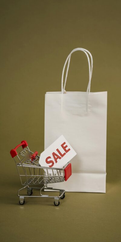 Photo by Sora Shimazaki A miniature shopping cart with a red sale tag beside a white shopping bag on a neutral background, perfect for retail concepts.