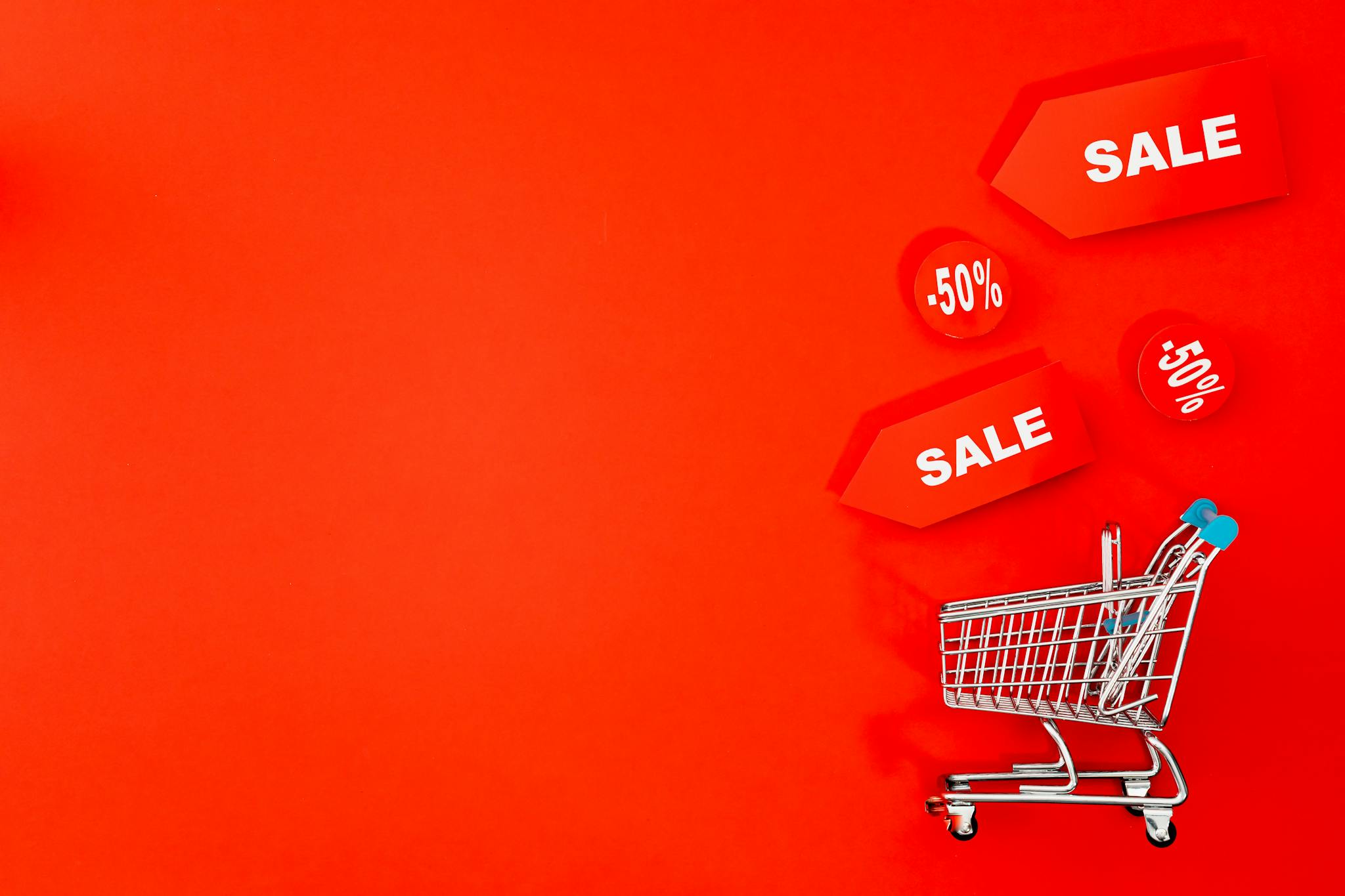 Shopping cart with sale tags on vibrant red backdrop promoting discounts.